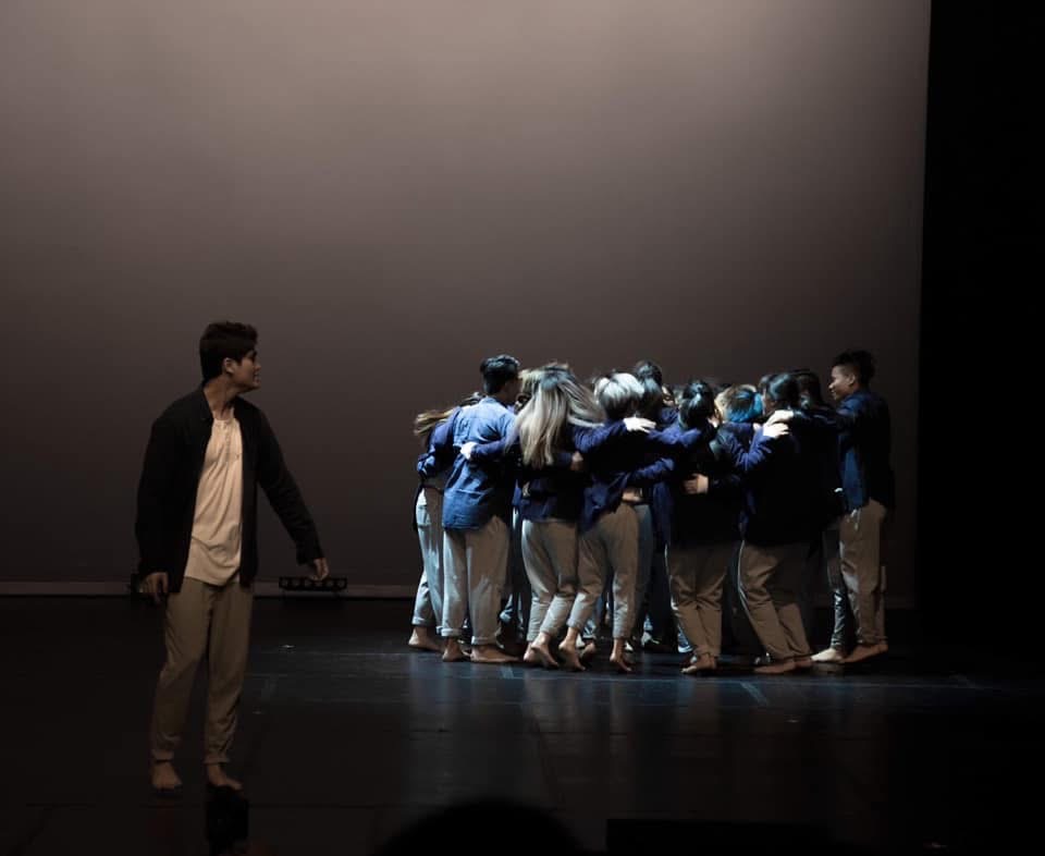 Jericho looking back at the I Swear I Lived ensemble huddled together on stage